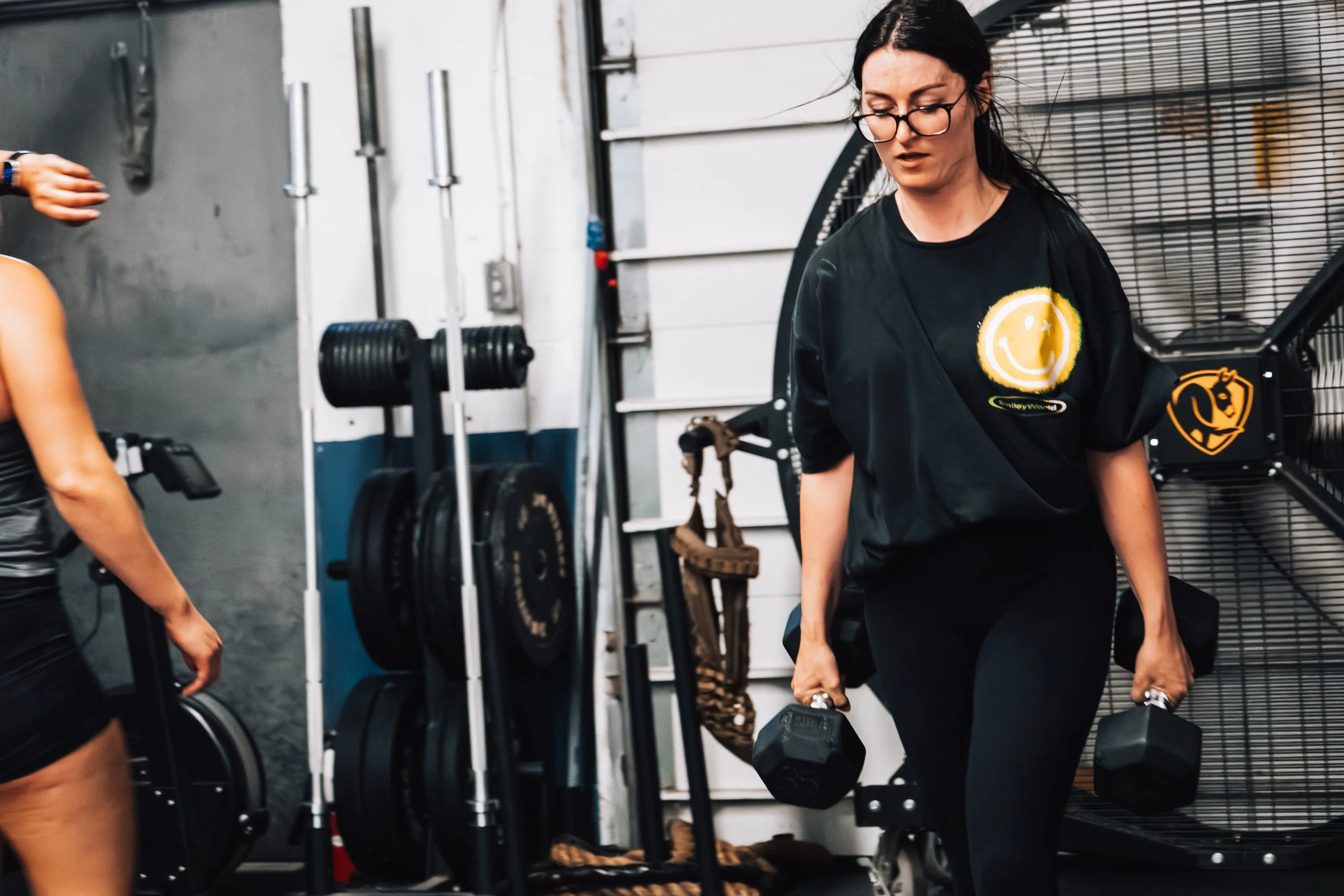A woman in workout clothes and glasses performing a walking lunge with dumbbells in a gym, with fitness equipment and weights around her.
