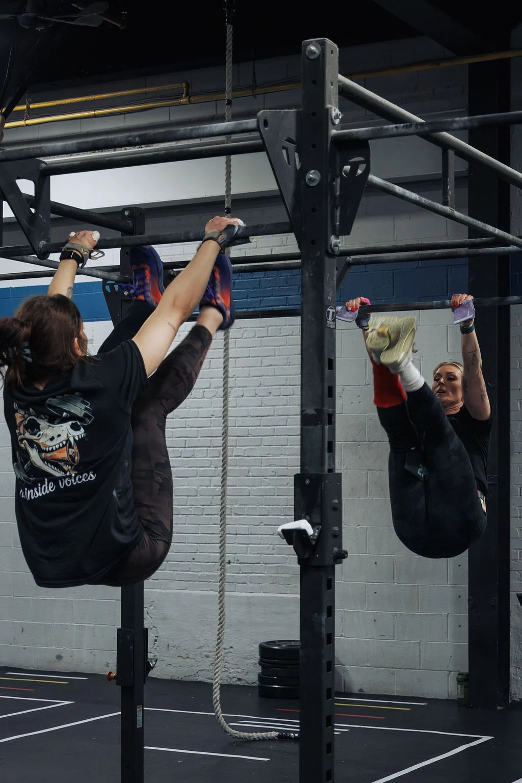 Two women participating in a gym workout hanging upside down from pull-up bars, one woman on each side of the structure, in an indoor gym space.