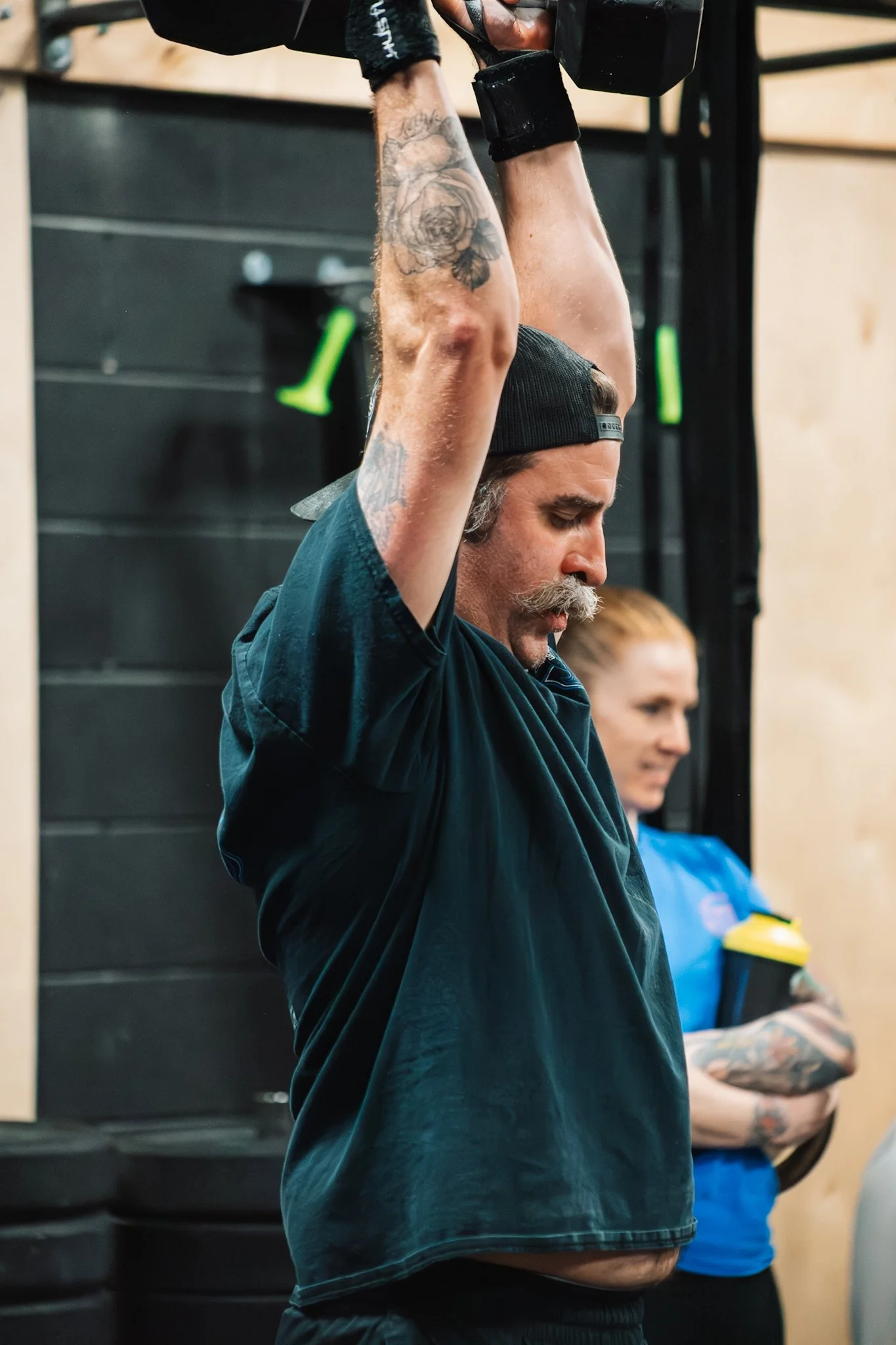 A man with tattoos on his arms wearing a black cap and shirt, lifting a black dumbbell overhead during a workout in a gym.