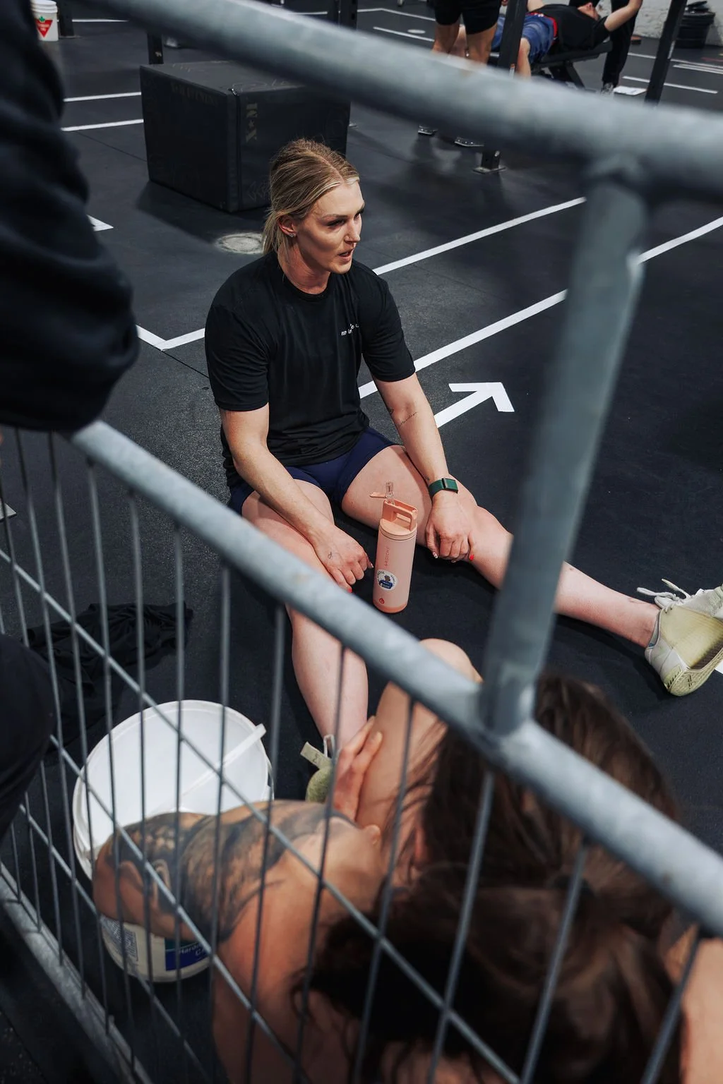 A woman sitting on the floor of a gym resting after exercise, with her leg stretched out and a water bottle by her side. Another person is lying down in front of her, and gym equipment can be seen around.