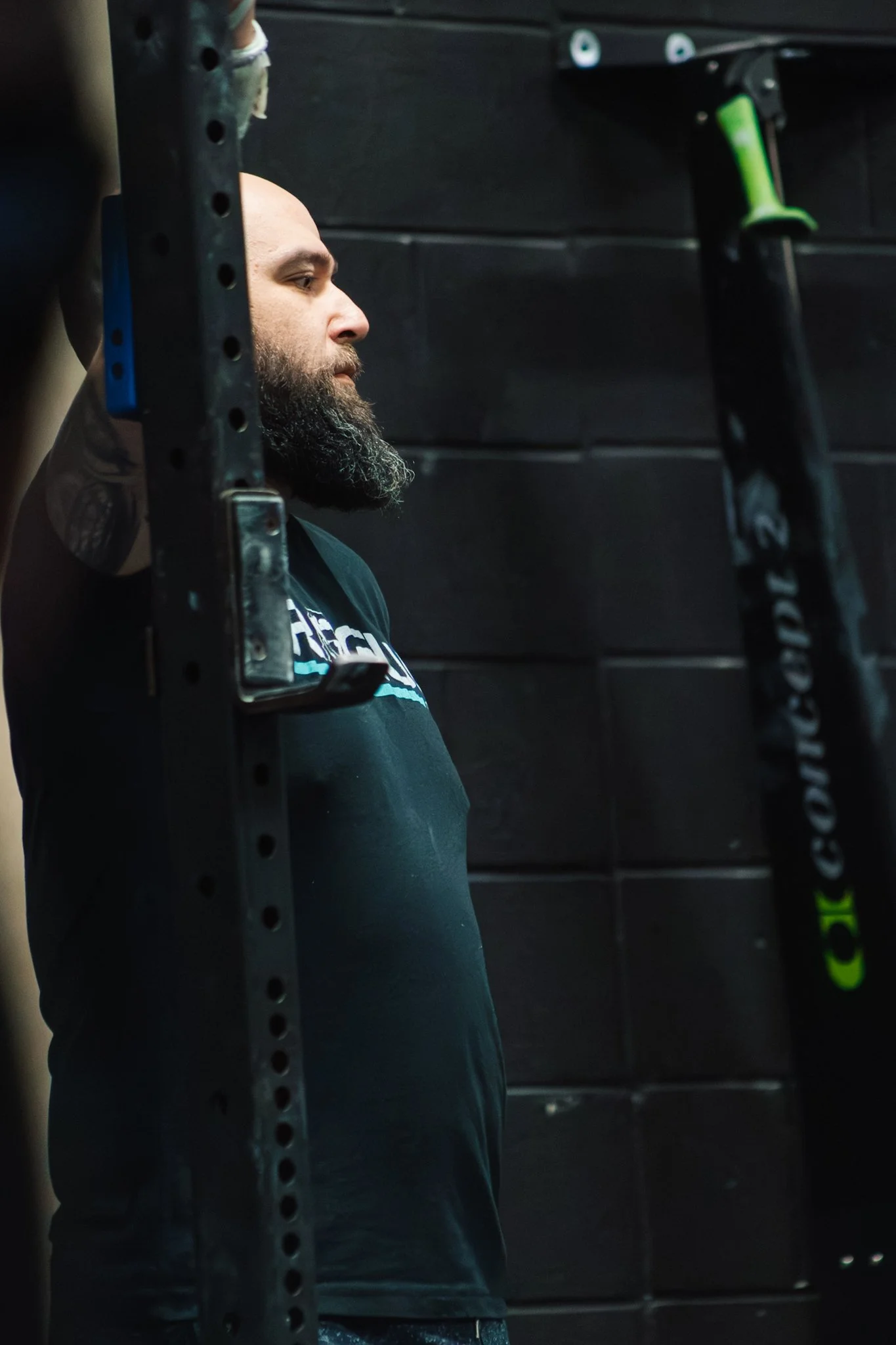 A man with a beard is leaning against a black wall with his eyes closed, appearing to rest or meditate in a gym or fitness environment, with gym equipment visible in the foreground and background.