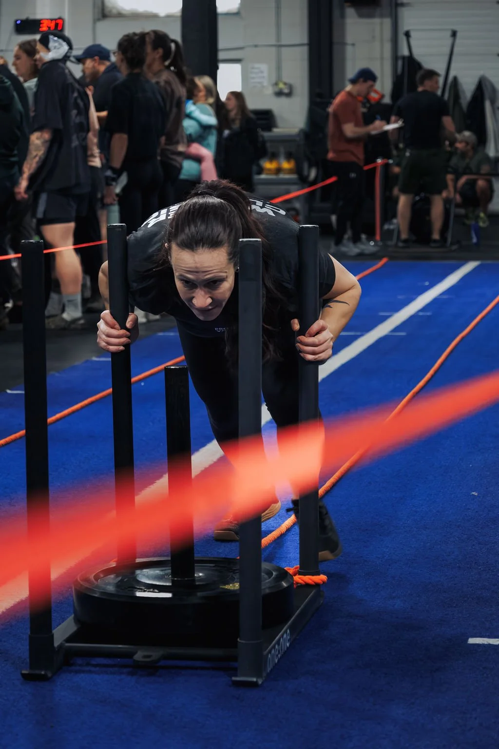 A woman pushing a weighted sled during a fitness competition or workout session inside a gym, with people standing and observing in the background.