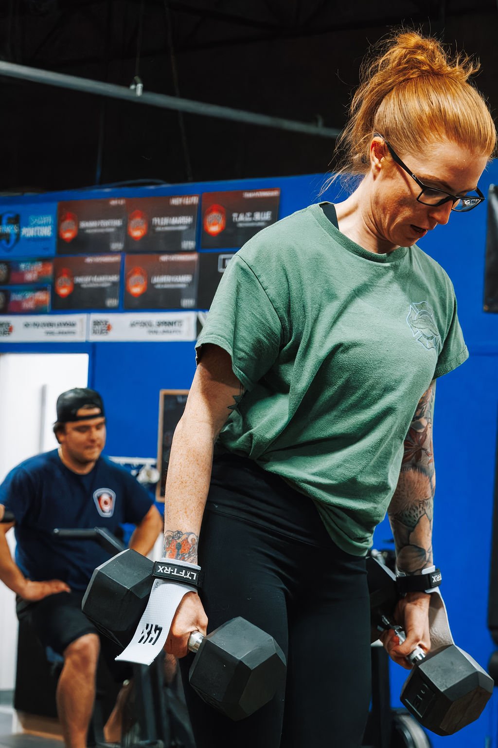 Woman with red hair, glasses, and tattoos lifting dumbbells in a gym. Man sitting in background in a blue T-shirt and black shorts.