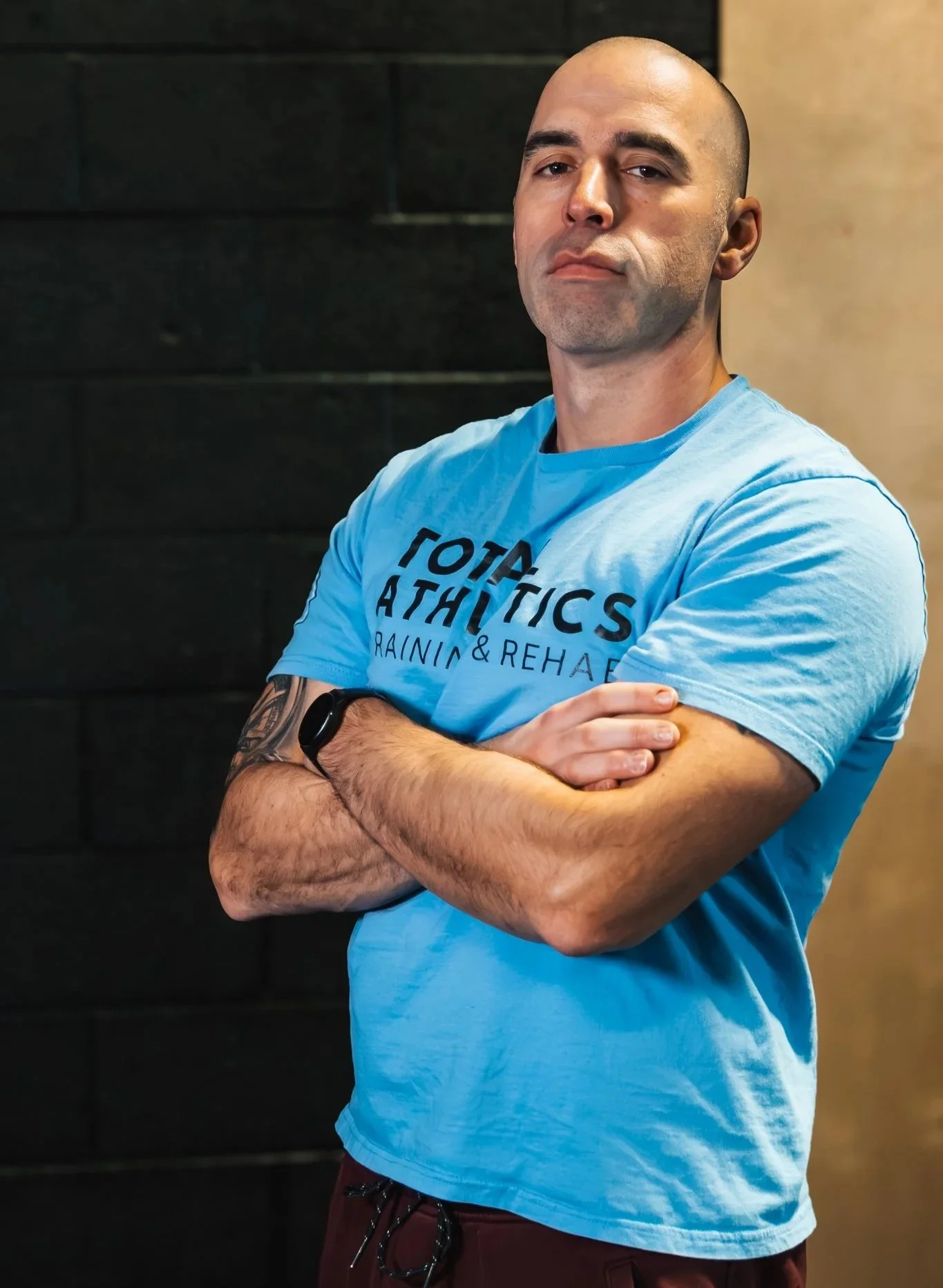 A man with a shaved head and a beard, smiling, wearing a black hoodie with blue and white logo text that reads "TOTAL ATHLETICS," standing with arms crossed against a gym background.