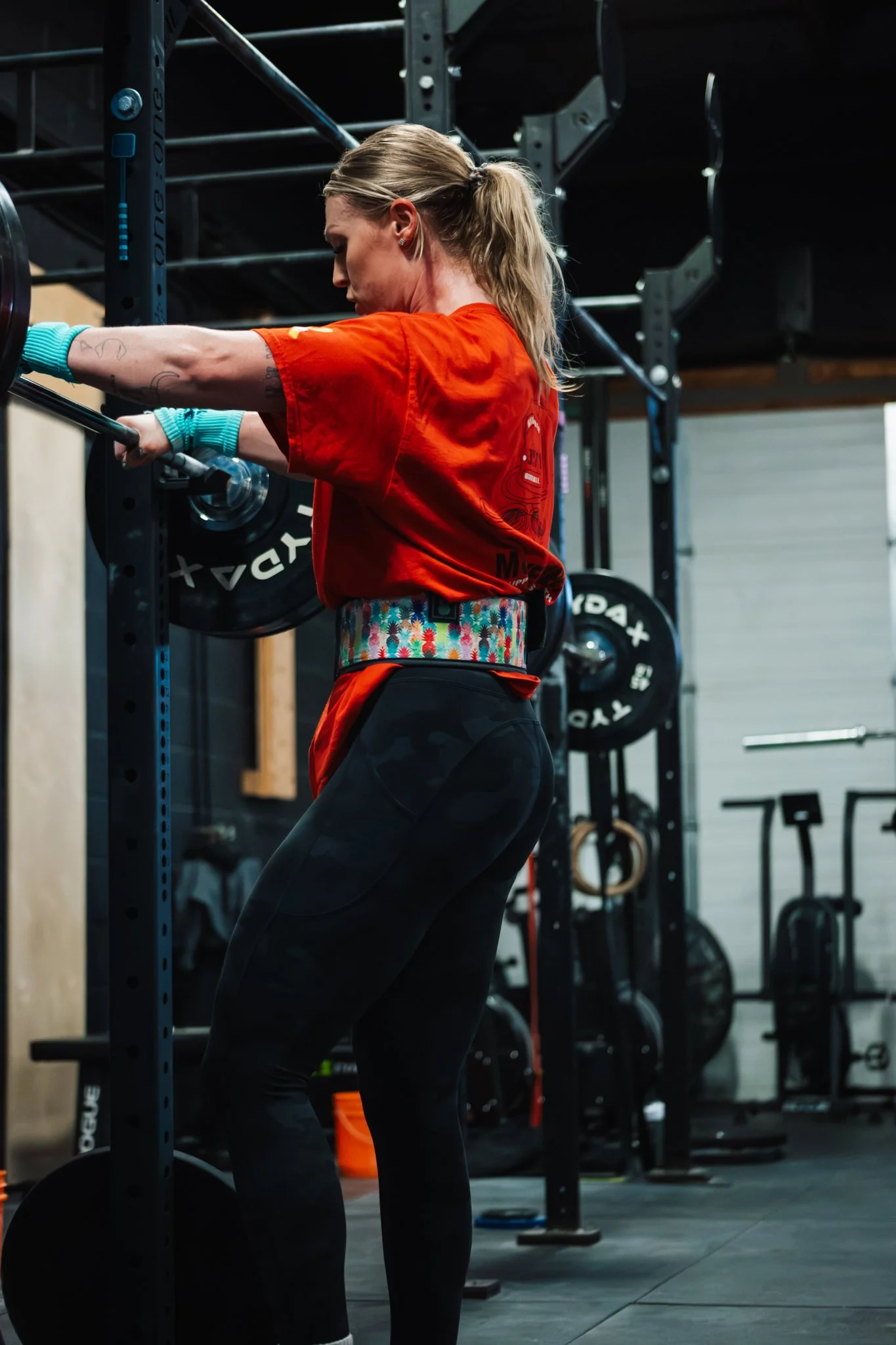 A woman with blond hair in a ponytail wearing a red T-shirt, black athletic leggings, teal wristbands, and a colorful weightlifting belt, stands and lifts a barbell in a gym.
