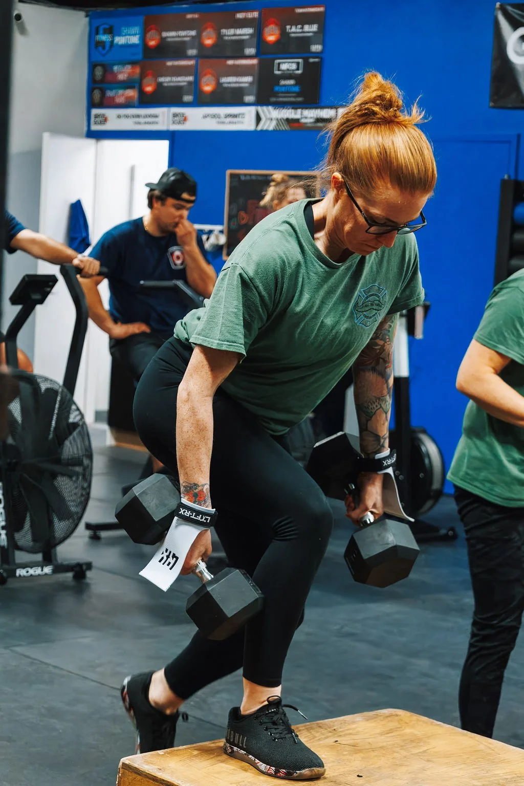 Woman with red hair, glasses, and tattoos wearing green shirt and black leggings, performing a weighted step-up exercise on a wooden box at a gym.