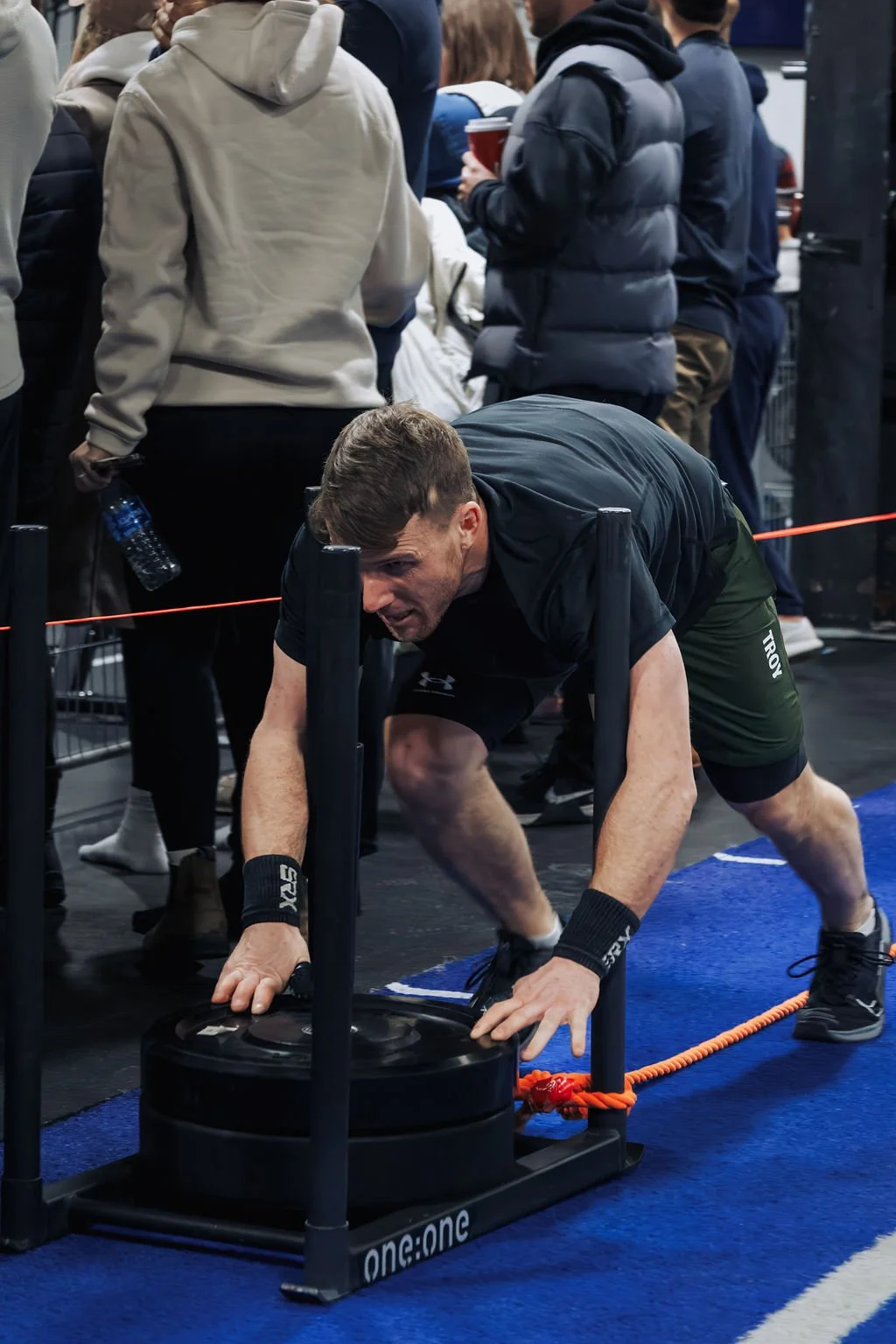A man in athletic clothing is intense and focused while performing a workout with a sled pushing exercise in a gym surrounded by other people.