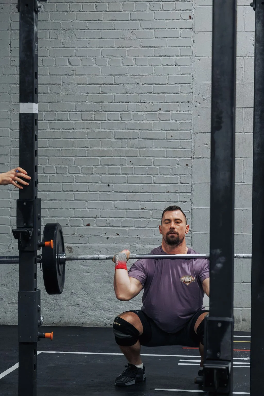Man performing a barbell squat in a gym, wearing knee sleeves and a gray T-shirt, with a secure grip on the barbell at shoulders level.