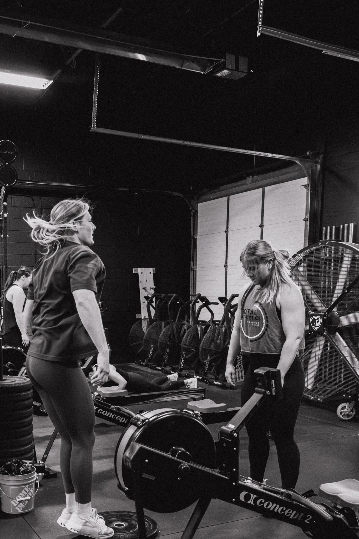 Two women working out with rowing machines in a gym, with other gym equipment in the background.