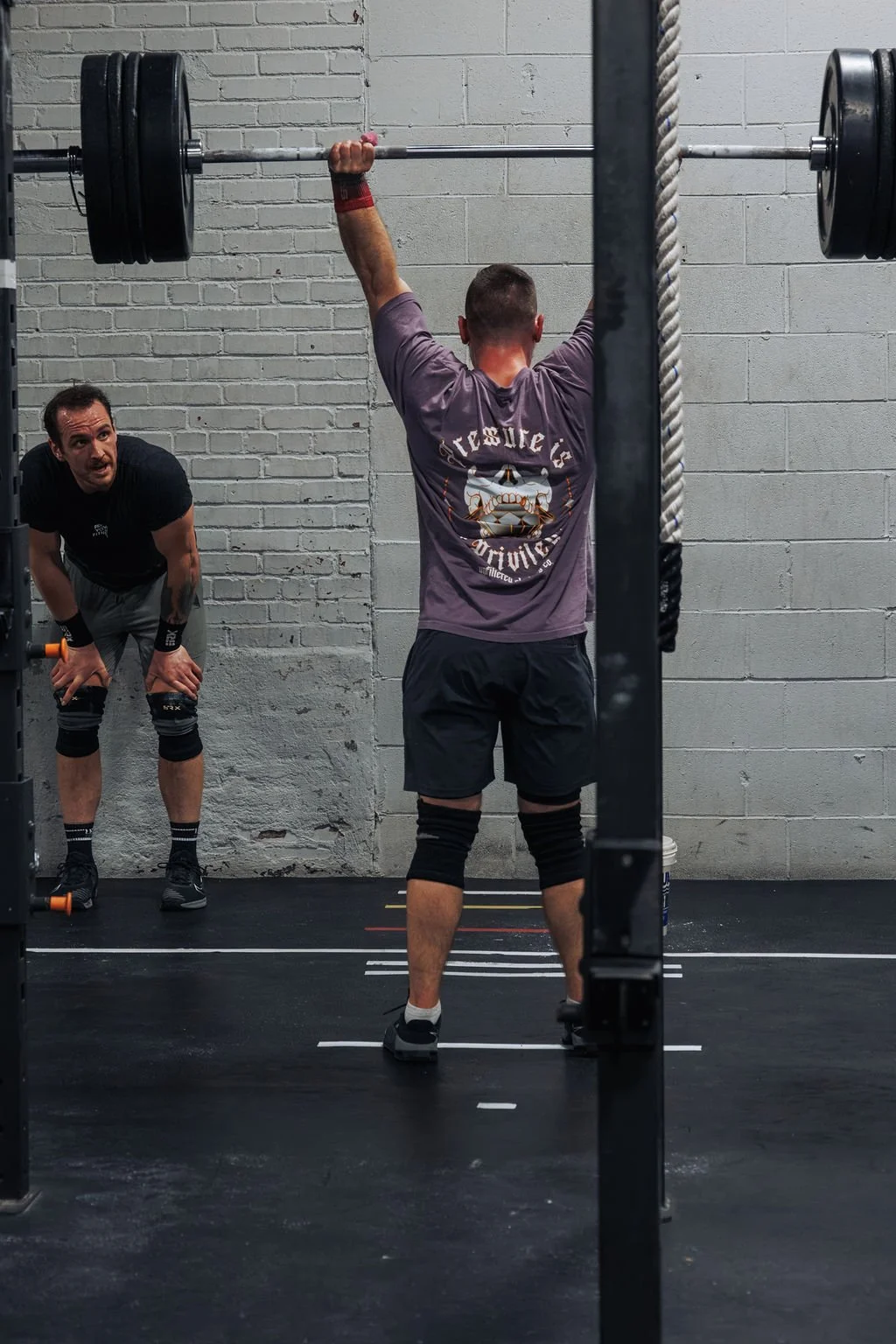 A man standing with his back to the camera, lifting a barbell overhead in a gym, while another man watches, leaning forward with hands on his knees.