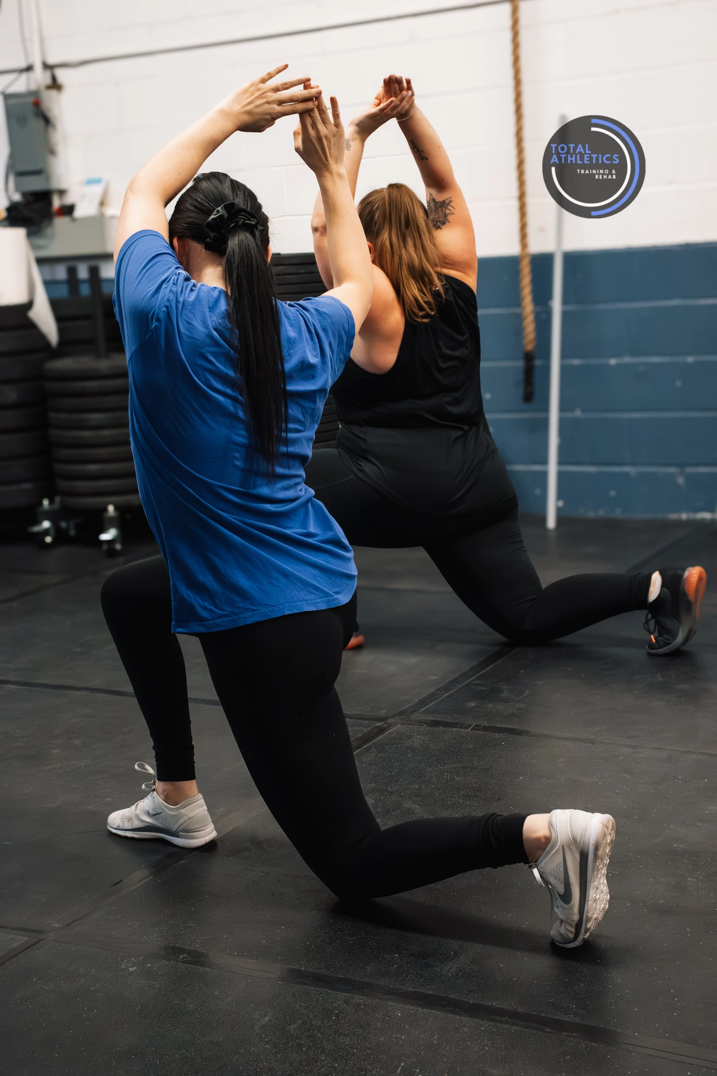 Two women performing a yoga pose in a gym, one in a blue shirt and black leggings, and the other in a black top and leggings, with a Total Athletics logo visible on the wall.