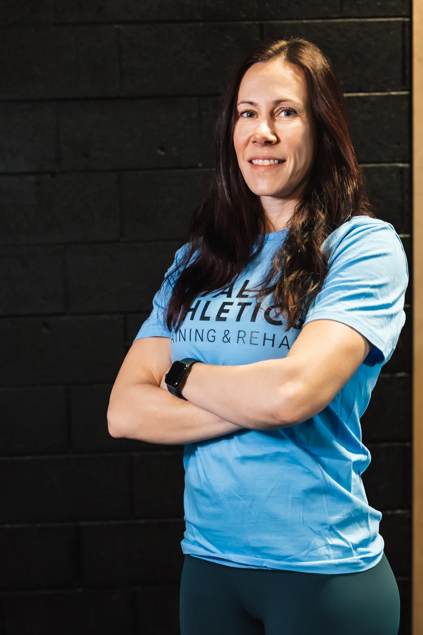 A woman with long dark hair smiling, wearing a blue athletic shirt and black leggings, standing cross-armed against a black wall.