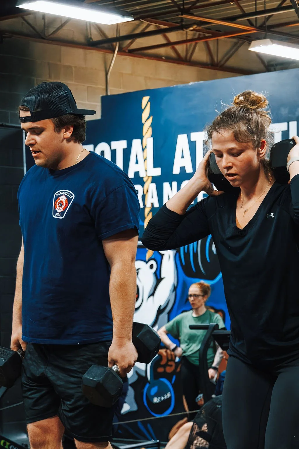 Two people lifting dumbbells at a gym, with a woman holding a weight on her shoulder and a man holding a weight in his hand, all in front of a blue wall with the text 'Total Attack' and a bear logo.