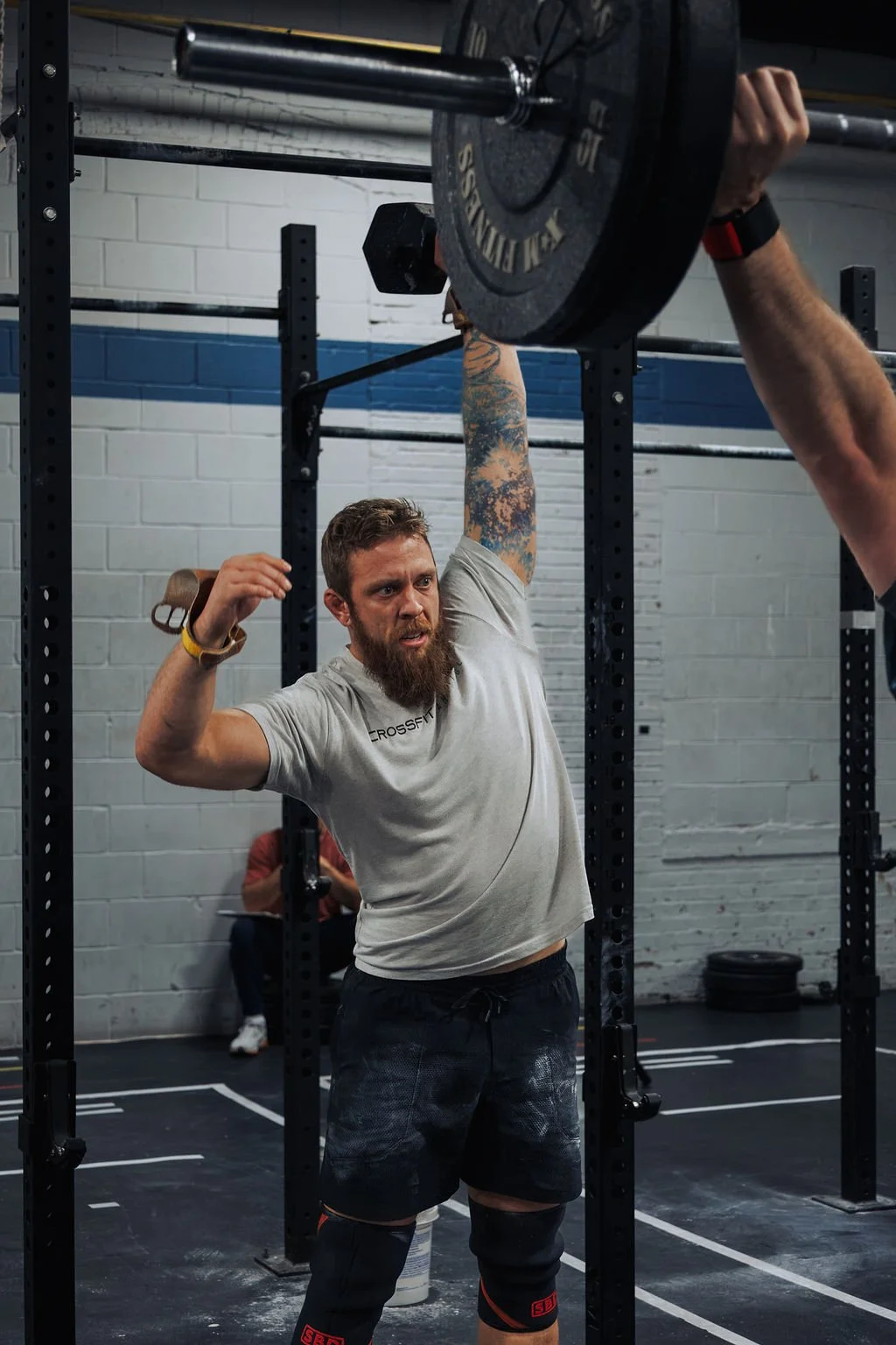 A man with a beard and tattoos on his arm is lifting a barbell overhead in a gym. He is wearing a gray T-shirt, black shorts, and knee sleeves. The gym has a black metal rack and a white brick wall in the background.