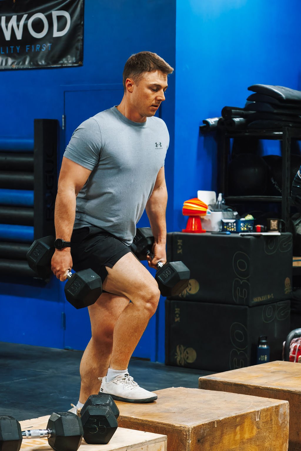 A man in a gray athletic shirt and black shorts is performing a workout on a plyometric box, holding dumbbells in each hand at a gym with blue walls and fitness equipment.