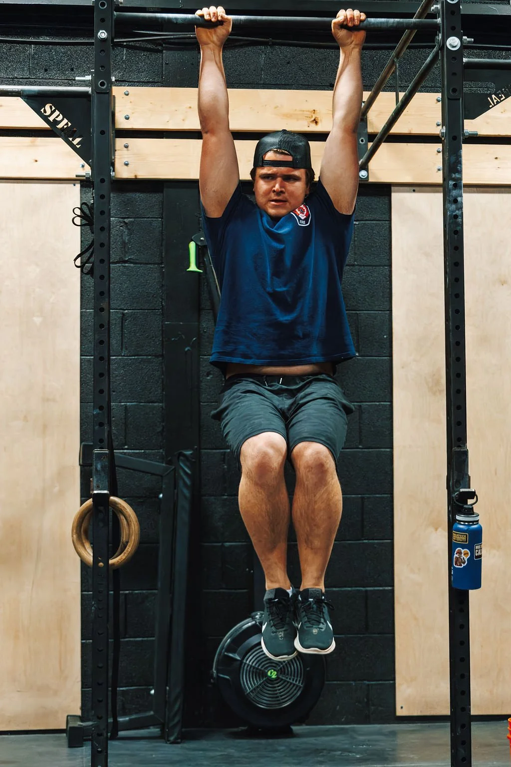 Man hanging from pull-up bars in gym, wearing black cap, navy shirt, and gray shorts.
