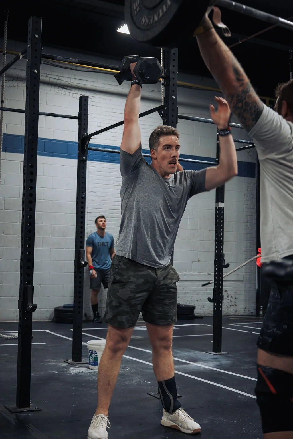 A man lifting a dumbbell overhead during a workout at a gym, with another person assisting or spotting him. A third person stands in the background near a wall.