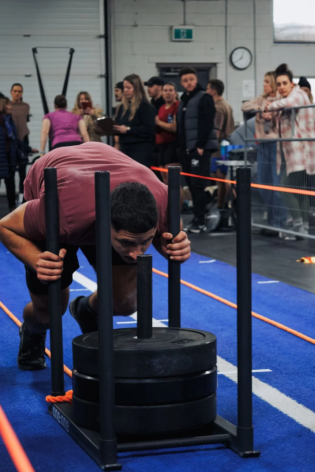 A man in a maroon shirt pushing a weight sled during a workout at a gym, with a crowd of people watching in the background.