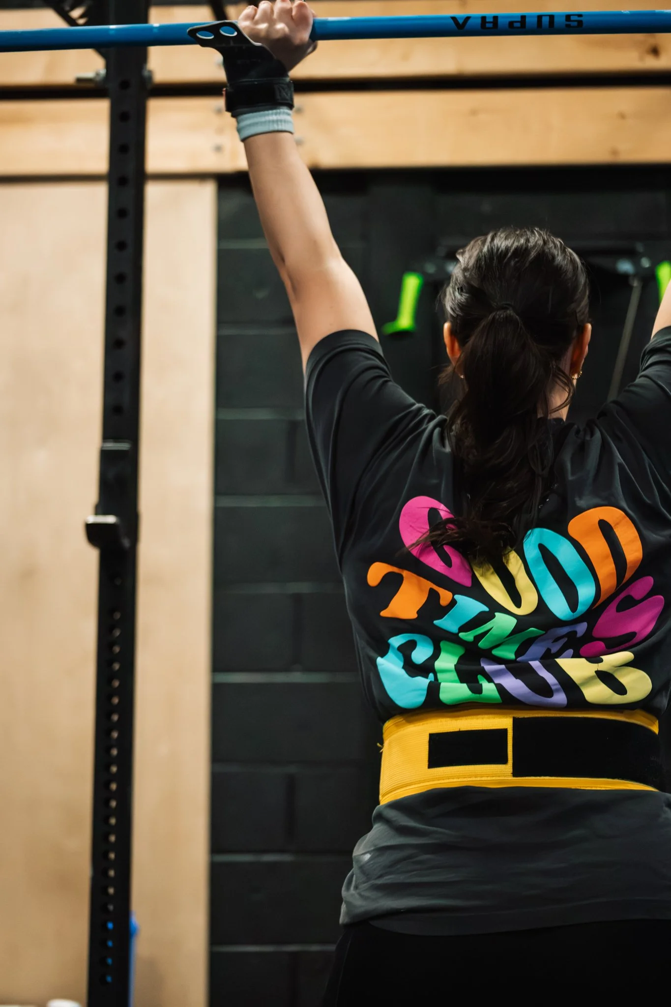 A woman with dark hair tied in a ponytail, wearing a black T-shirt with colorful text, is seen lifting a blue weight bar overhead in a gym.