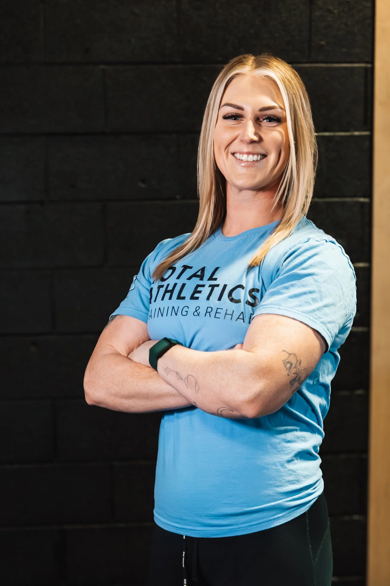 A smiling woman with long blonde hair, wearing a blue T-shirt with the text "TOTAL ATHLETICS TRAINING & REHAB," standing with arms crossed against a black brick wall.