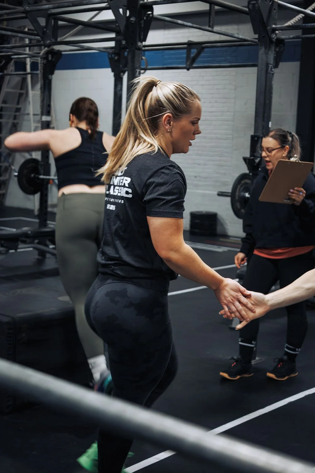 A woman with blonde hair in a ponytail, wearing a black t-shirt and camouflage leggings, is holding hands with another person in a gym environment. There is a woman with glasses holding a clipboard and a woman with a barbell in the background.