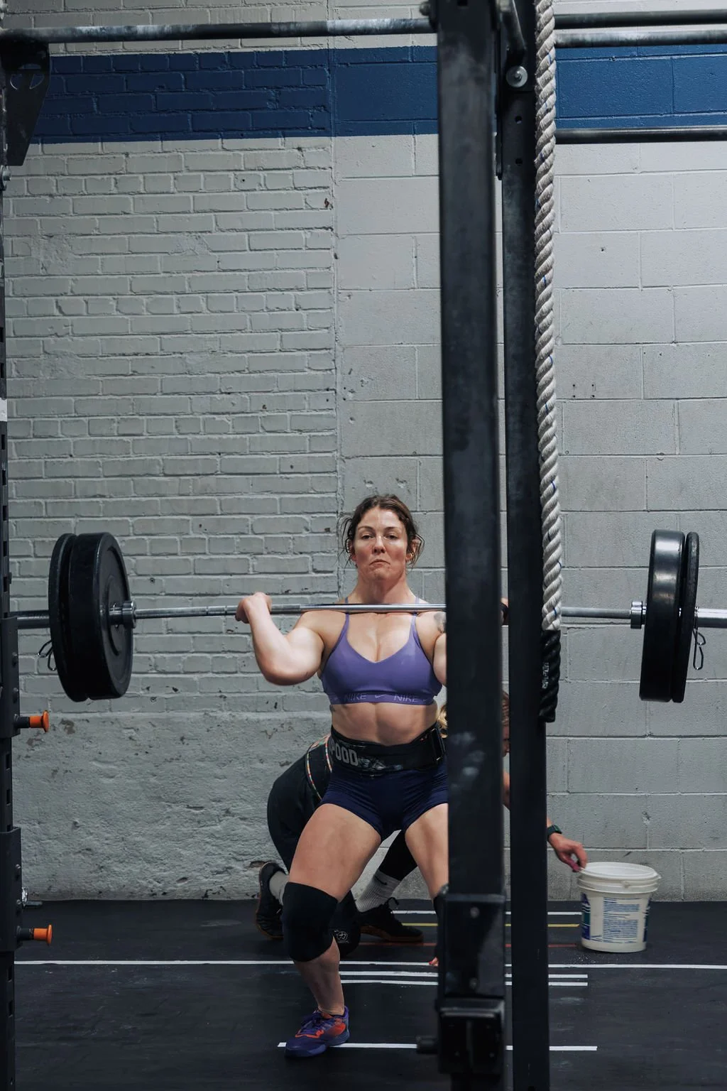Female weightlifter in a purple sports bra and black shorts performing a squat with a barbell loaded with weights in a gym with gray brick walls.