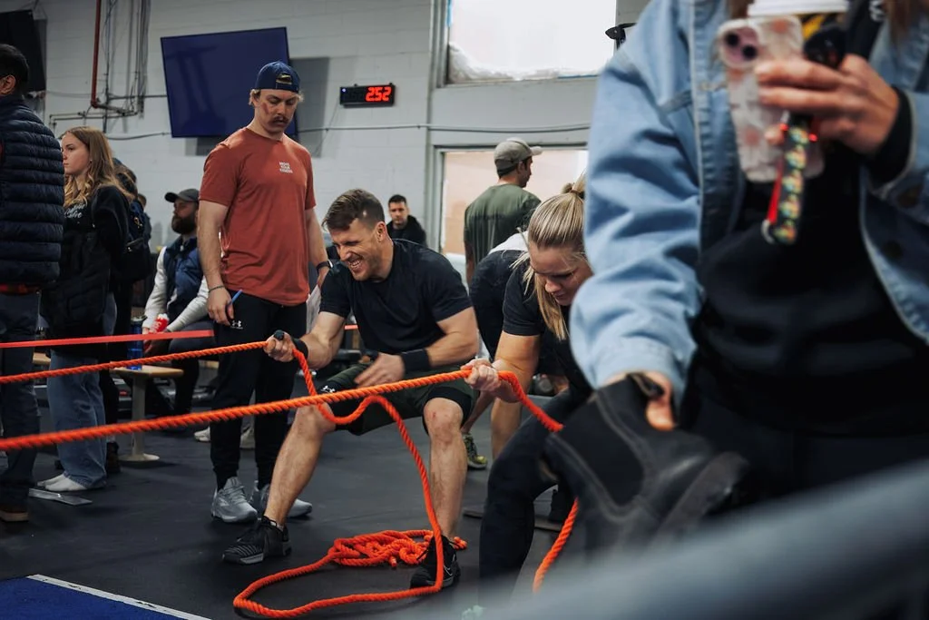 People participating in a tug-of-war competition indoors, with some showing effort or amusement. Several onlookers are in the background, and a digital timer reads 2:52.