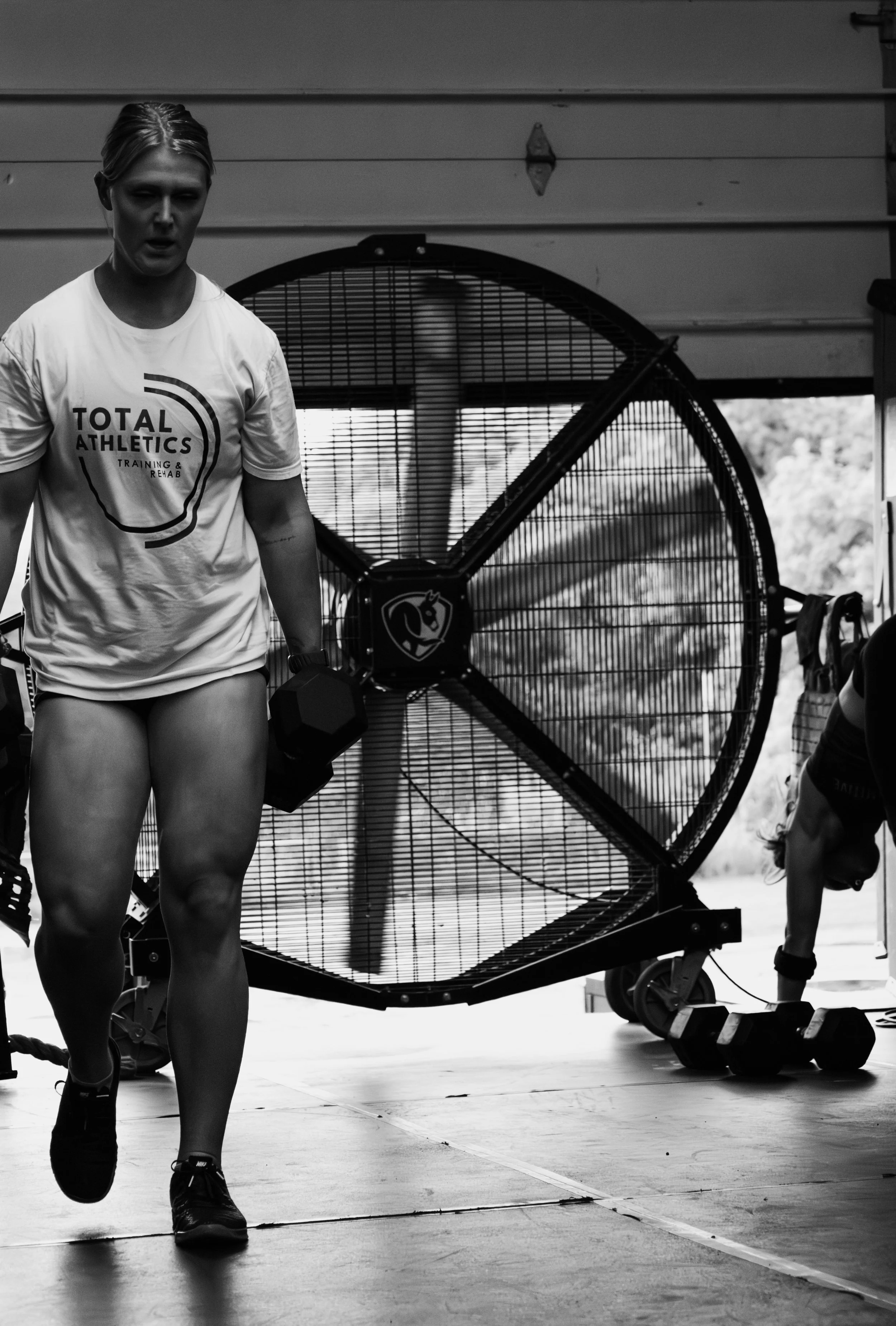 A woman in a white T-shirt holding a dumbbell in an indoor gym with industrial equipment and a large fan in the background.
