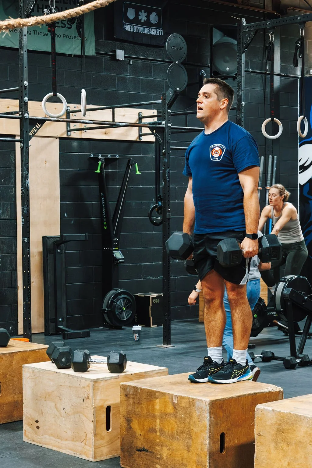 A man in a blue T-shirt and black shorts holding dumbbells during a workout in a gym, with women exercising in the background and various gym equipment around.