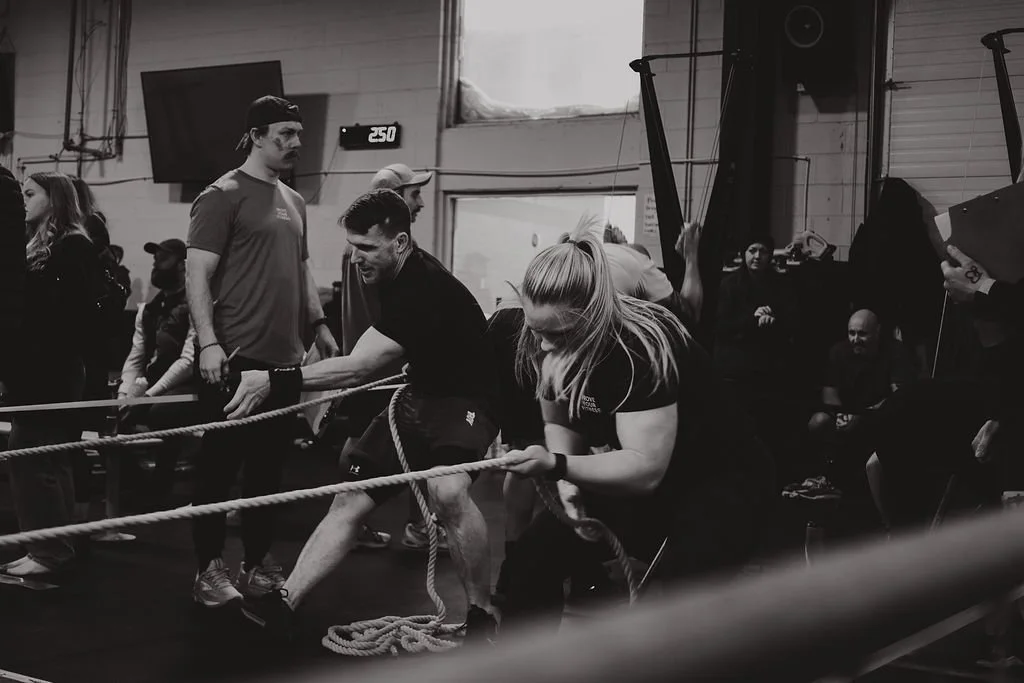 People participating in a ropes challenge in a gym, with some pulling a rope and others watching. The scene is indoors with fitness equipment and a digital clock showing 2:50.