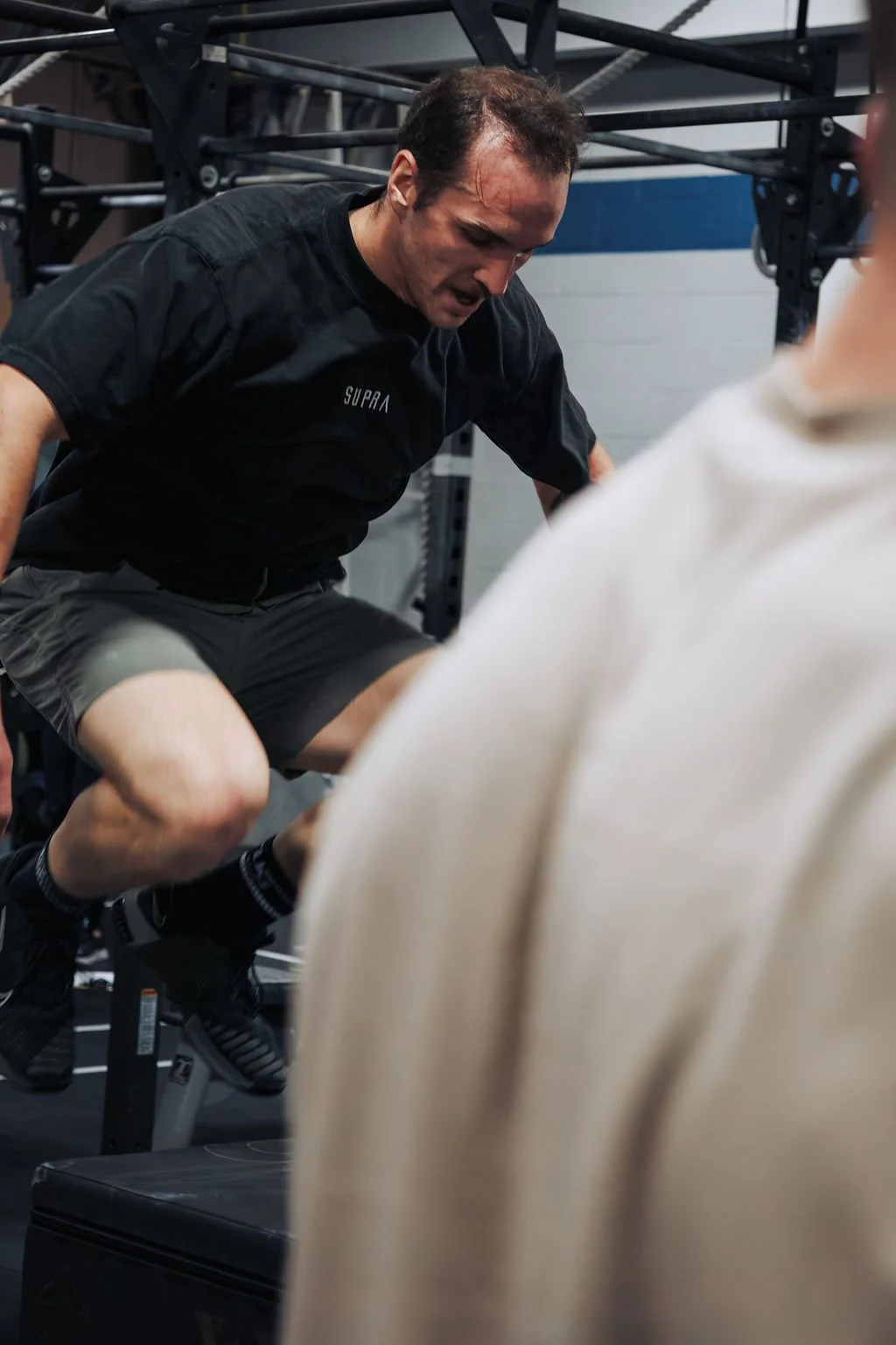 A man wearing a black t-shirt and gray shorts doing a step-up exercise with a weighted vest in a gym.