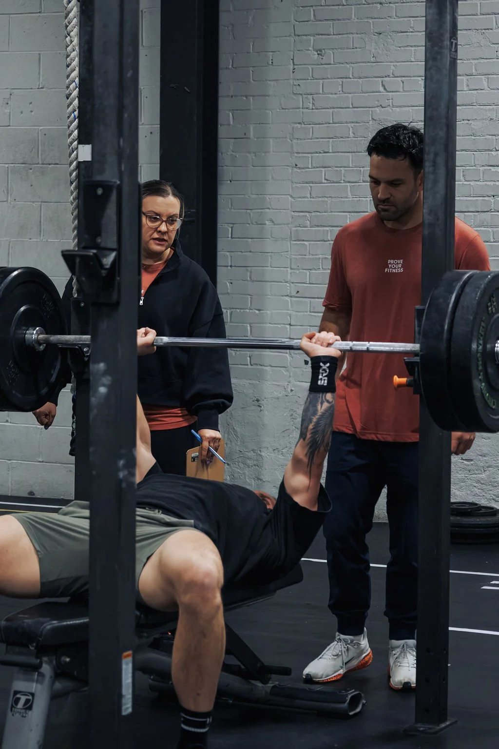 Man lying on a bench press with a barbell, while a woman and a man spot him in a gym with a gray brick wall.