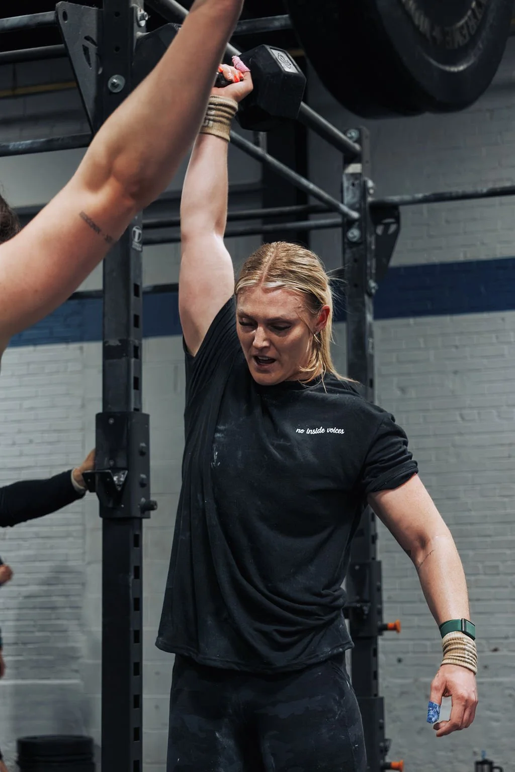A woman with blonde hair and a black T-shirt is lifting a dumbbell overhead in a gym, appearing to be exhausted after a workout.