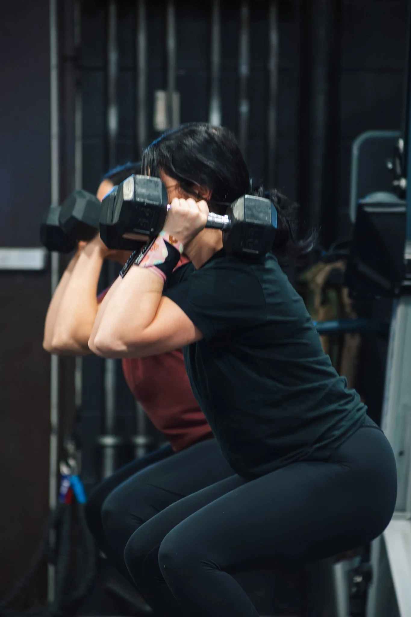 Two women engaging in a workout, performing squats with dumbbells at a gym.