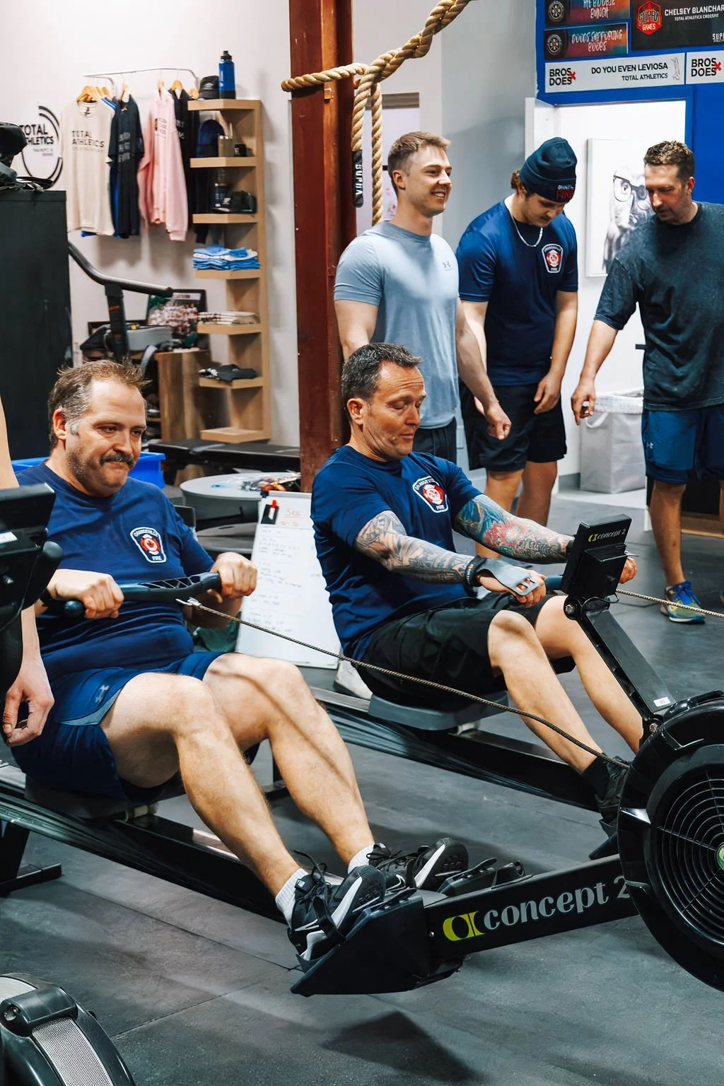 Group of men working out on rowing machines in a gym, with some standing and watching.