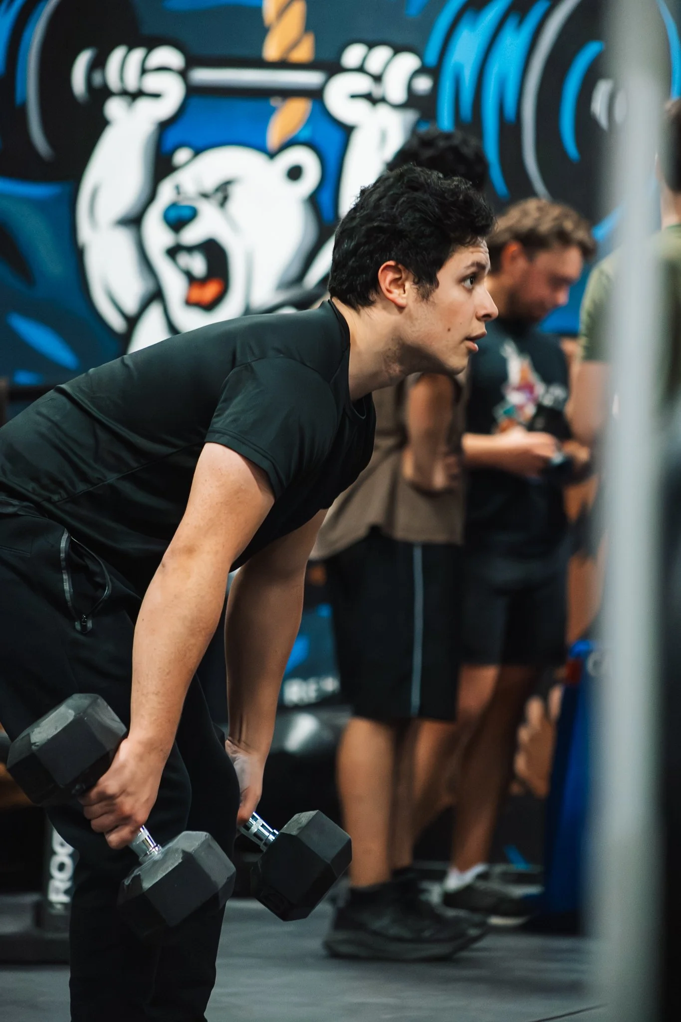 A young man in black workout clothes is lifting dumbbells in a gym with other people in the background, and a bear graphic on the wall.