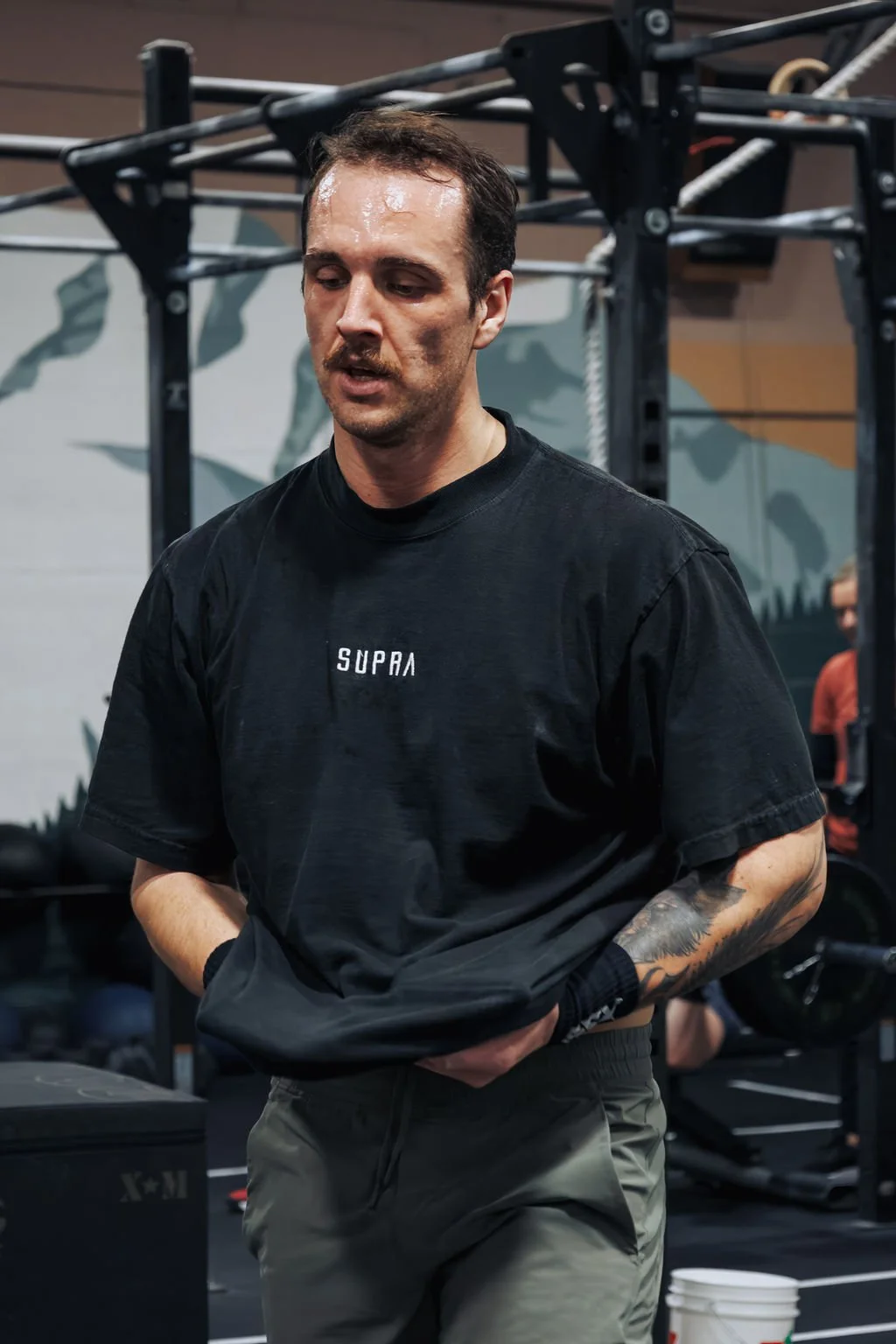 A man with a mustache and tattoo on his arm, wearing a black "SUPRA" t-shirt and gray pants, standing in a gym with workout equipment and a mural in the background.