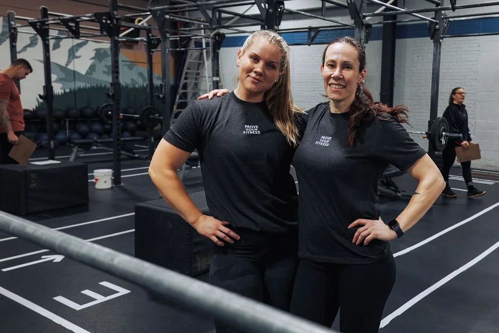 Two women standing together in a gym, wearing black workout shirts, smiling, with exercise equipment in the background.