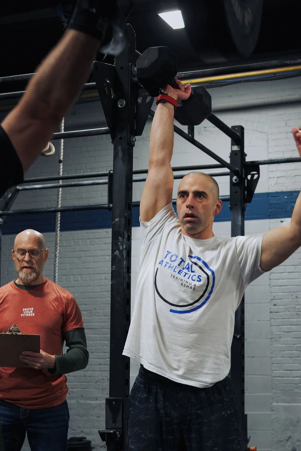 Man lifting a dumbbell overhead during workout at gym, with trainer holding clipboard observing