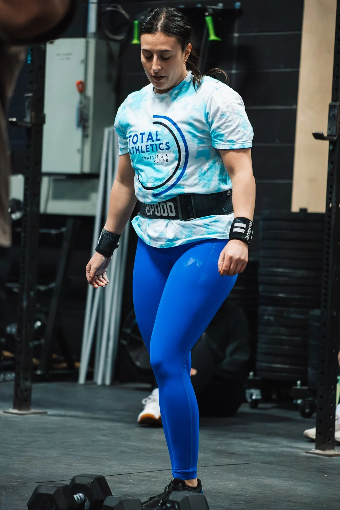A woman in blue athletic leggings, a white T-shirt with 'Total Athletics' on it, and a black weightlifting belt in a gym setting, preparing to lift weights.