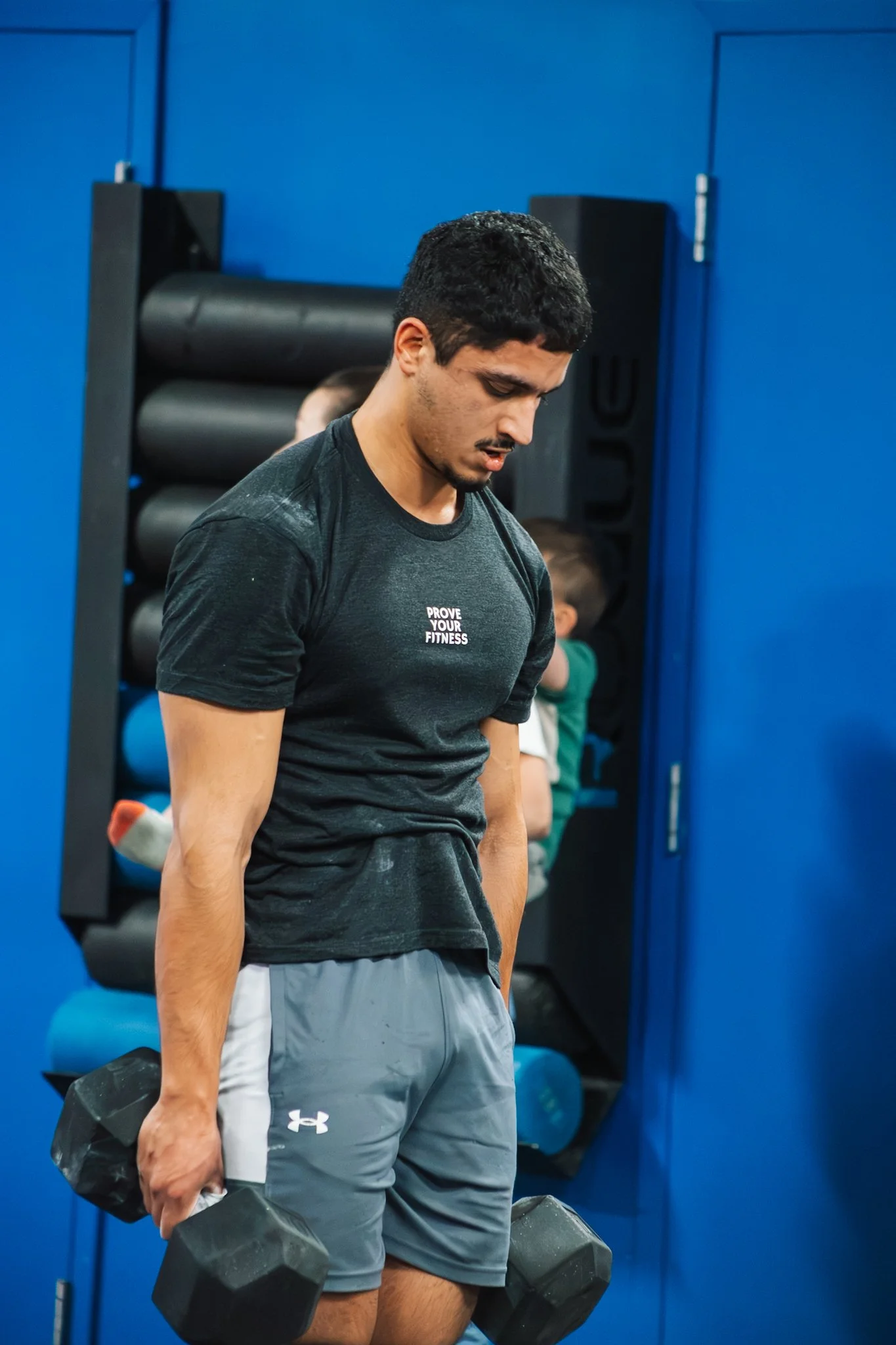 Man in black athletic shirt and gray shorts lifting dumbbells in gym, with focused expression and other people in background.