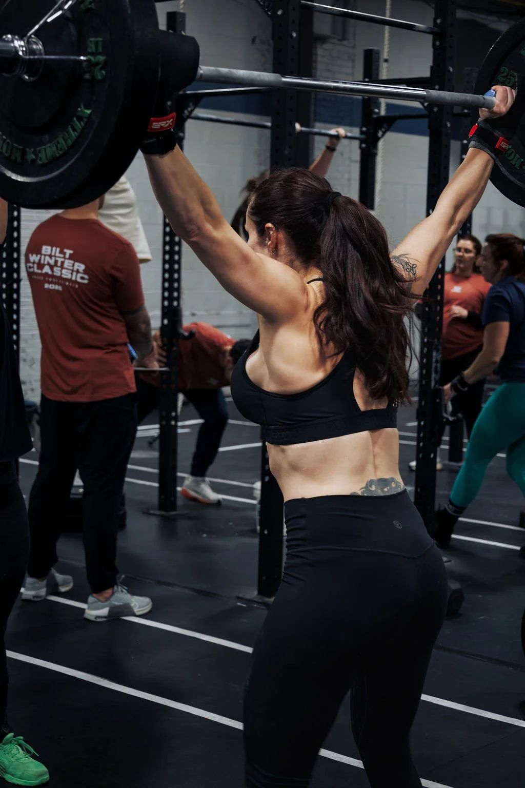 A woman lifting a barbell overhead during a workout at a gym, surrounded by other people exercising.