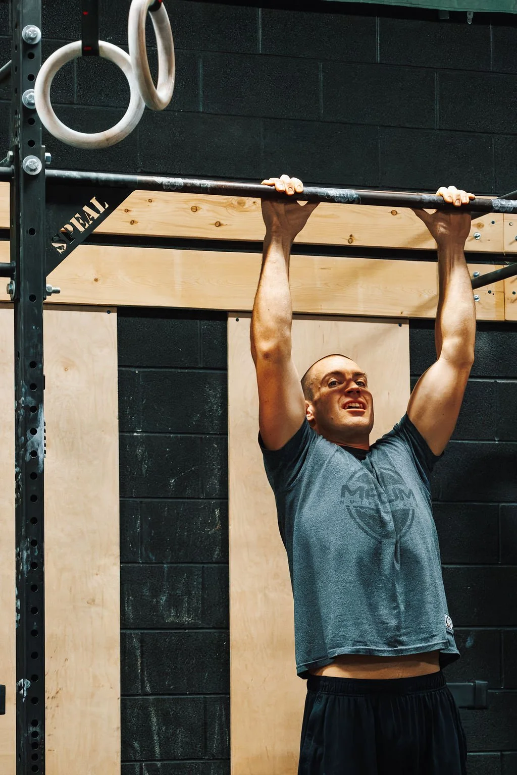 A man hanging from a pull-up bar in a gym, with gymnastic rings hanging nearby.