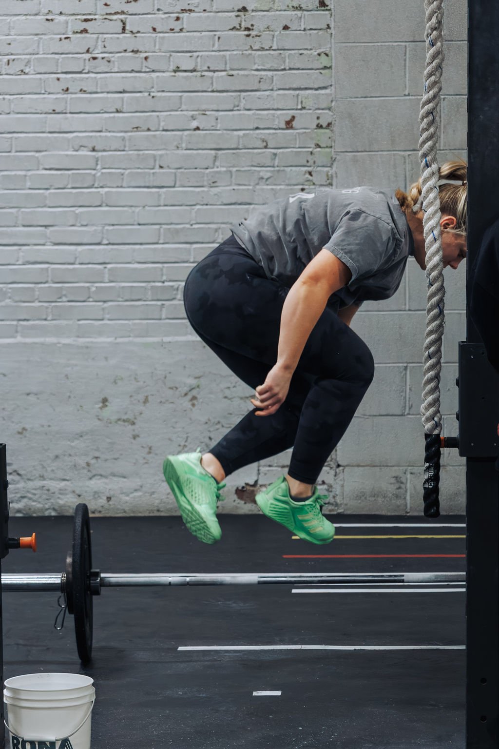 A woman mid-air during a workout, doing a pull-up, inside a gym with a brick wall background, green athletic shoes, and a barbell on the floor.