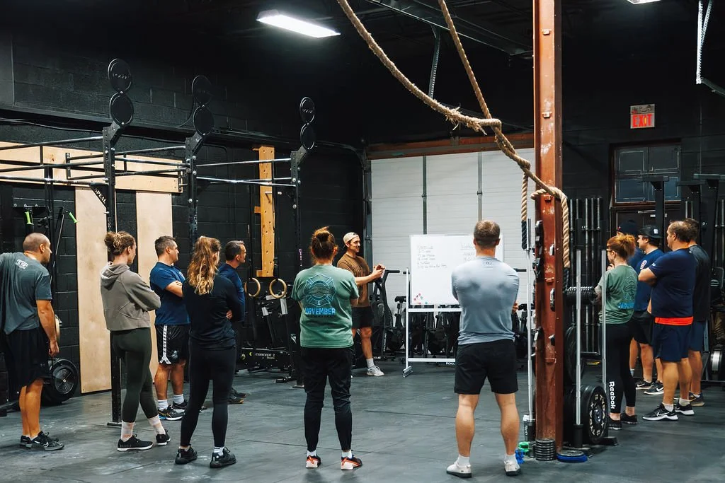 A group of people in a gym, standing in a semi-circle around a trainer who is giving instructions near a whiteboard.