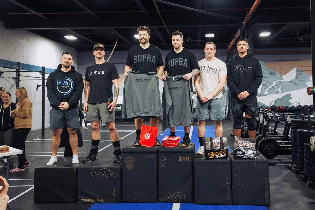 Six men standing on a winners' podium in a gym, holding sport jackets, with awards and gear displayed in front. People are in the background, and gym equipment surrounds them.