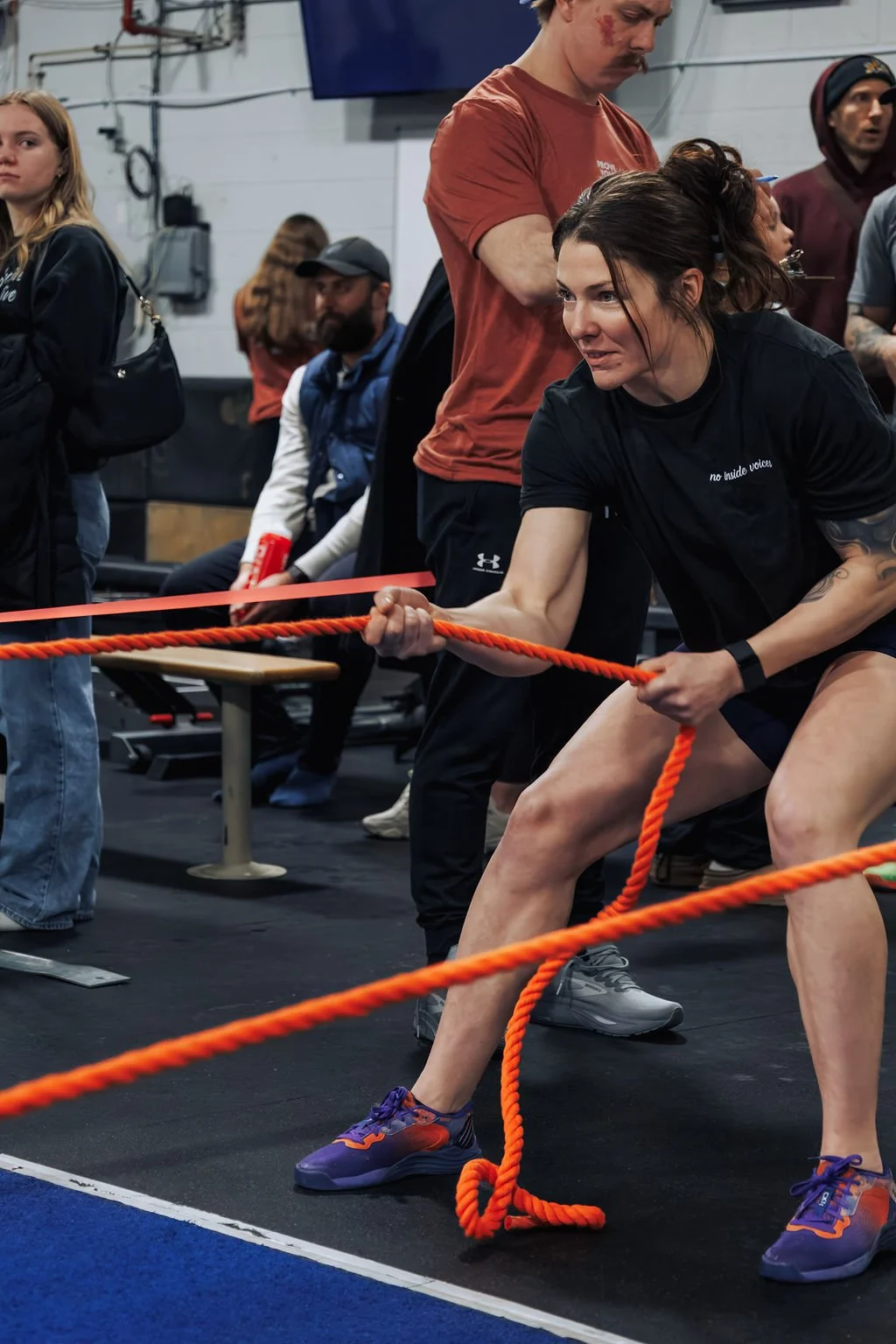 A woman with a tattoo on her right arm is participating in a strongman competition, pulling a thick orange rope with a determined expression. She is wearing a black t-shirt, black shorts, and purple athletic shoes. Several spectators and officials ar