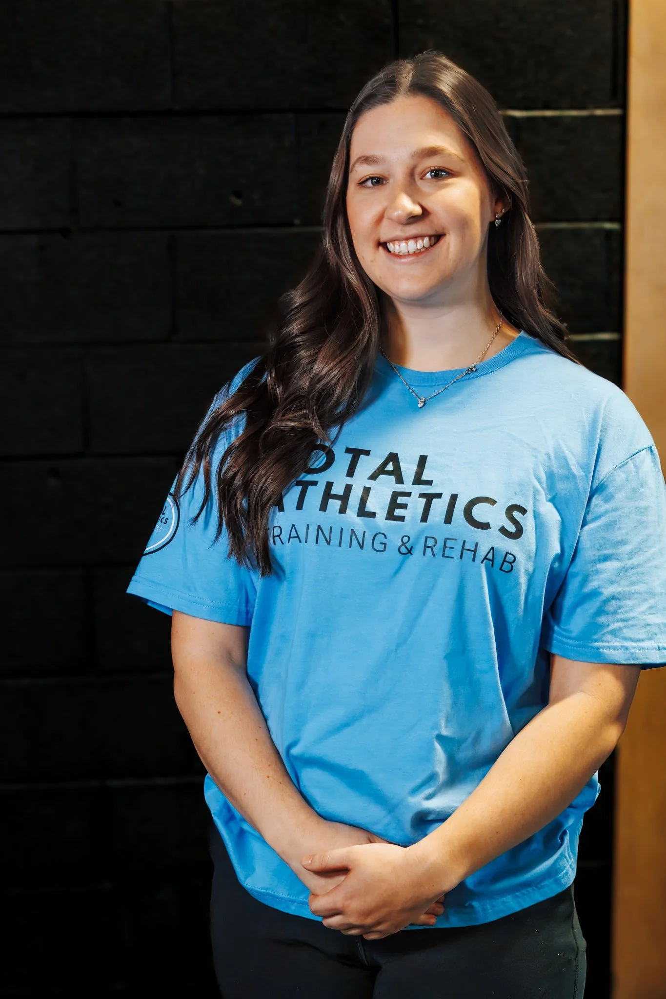 Young woman smiling, wearing a blue T-shirt with "Total Athletics Training & Rehab" written on it, standing against a dark background.