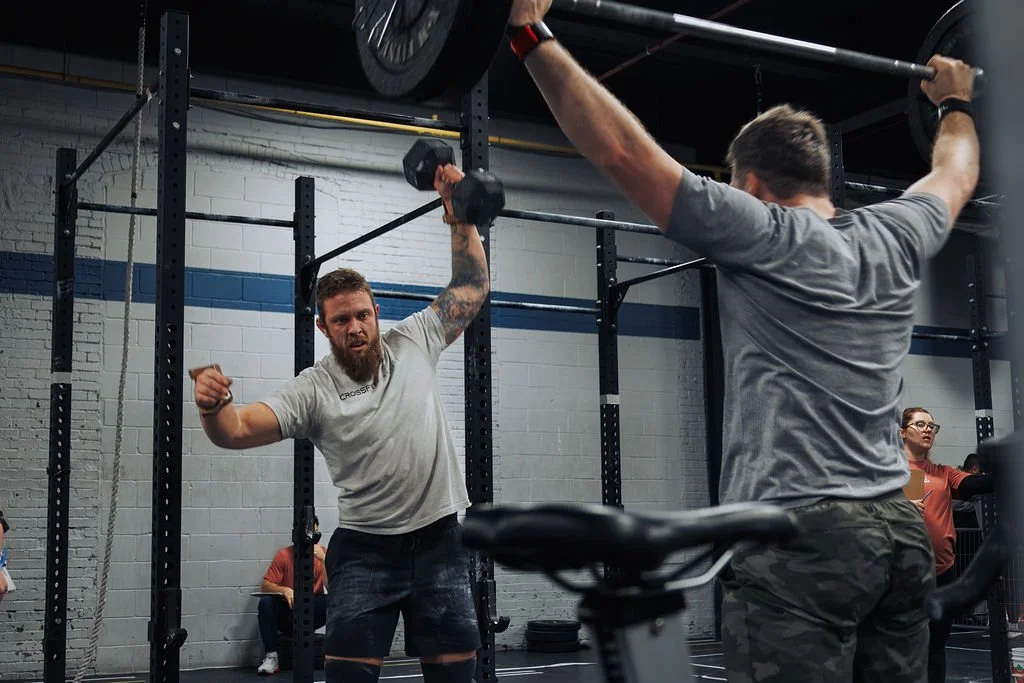 Two men working out in a gym. One man is lifting a barbell overhead, and the other man is standing nearby watching. There are other people in the background.