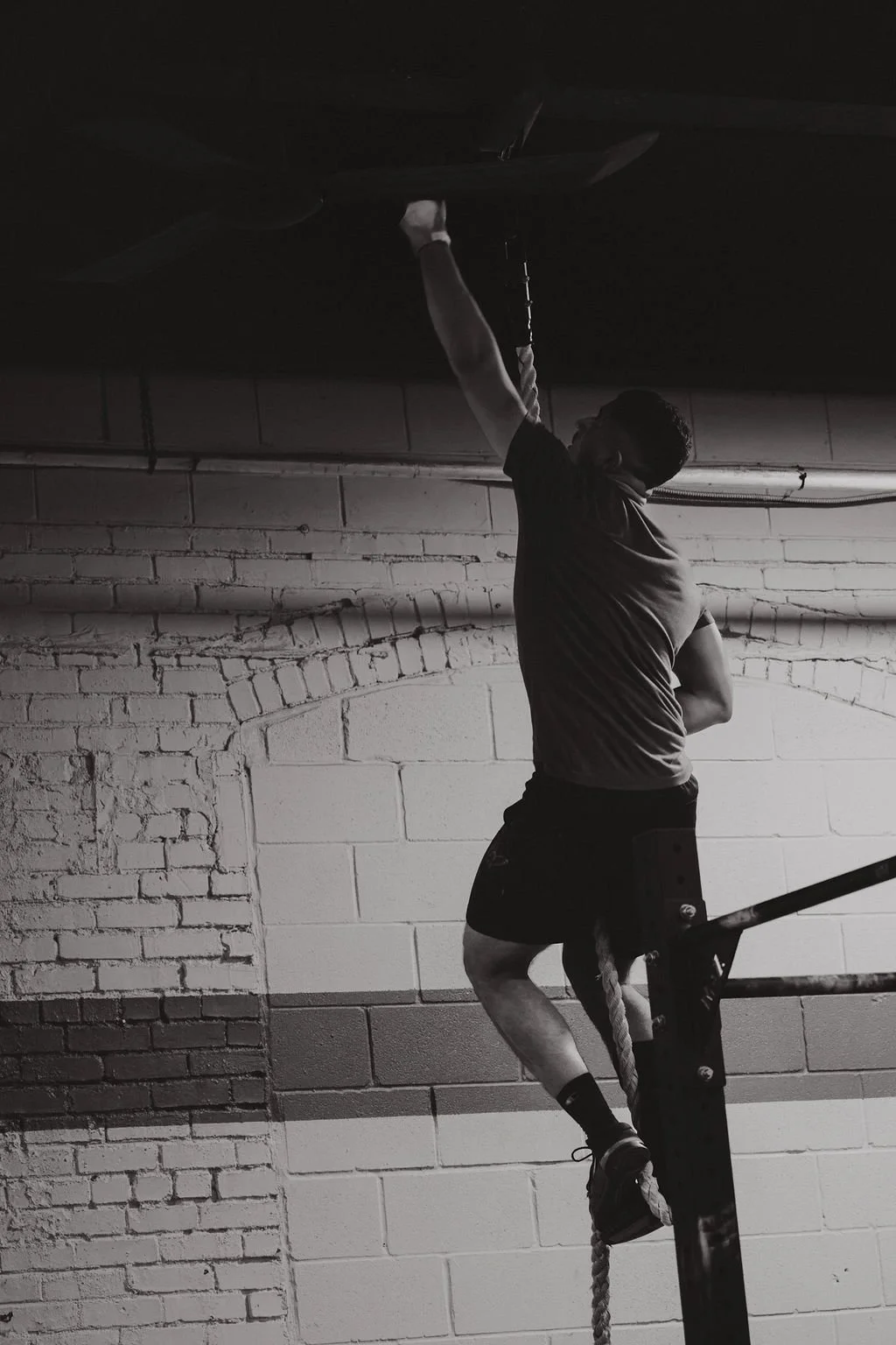 A person climbing a rope in an indoor gym, with a brick wall in the background, black and white photo.